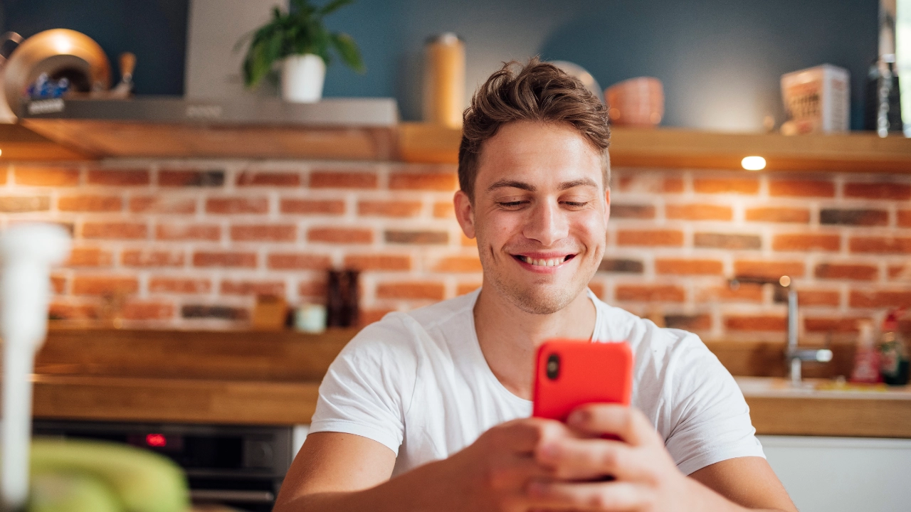 Ein junger Mann sitzt in der Küche und schaut in sein Handy.